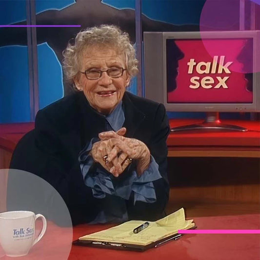 Sue Johanson smiling at her desk on the TV set of Talk Sex, with a notepad, pen, and coffee mug beside her.
